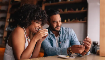 Woman holding a coffee mug while looking at man who’s showing her something on his phone.