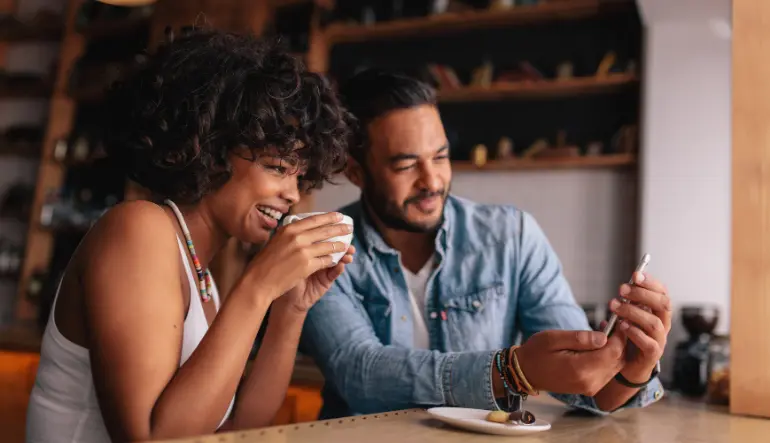 Woman holding a coffee mug while looking at man who’s showing her something on his phone.